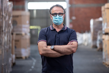 Uomo con occhiali neri e mascherina protettiva incrocia le braccia nel magazzino in cui lavora