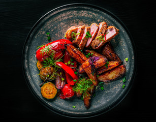 fried meat with potatoes, pepper, tomatoes, herbs and spices in plate on black wooden table background