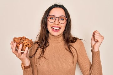 Beautiful woman with blue eyes holding bowl with healthy almonds over white background screaming proud and celebrating victory and success very excited, cheering emotion