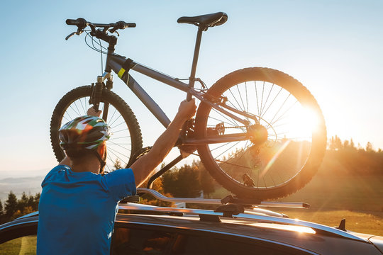 Young Man dresses modern cycling clothes and protective helmet installing his mountain bike on the car roof with sunset backlight. Active sporty people concept image.