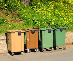 Plastic bins of different colors for garbage collection of different types in a Spanish street with contrast of a natural green background with flowers