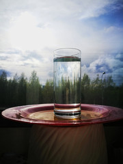 A transparent glass with clear water stands on a red-orange plate against the background of a window with a view of green trees and the sky in the clouds.