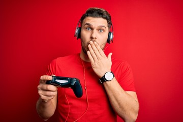 Young gamer man with blue eyes playing video games using gamepad joystick cover mouth with hand shocked with shame for mistake, expression of fear, scared in silence, secret concept