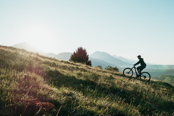 Athletic man pedalling an MTB E-bike up a steep grassy hill. Beautiful view of the mountains at...