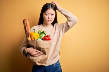 Young asian woman holding paper bag of fresh healthy groceries over yellow isolated background confuse and wondering about question. Uncertain with doubt, thinking with hand on head. Pensive concept.