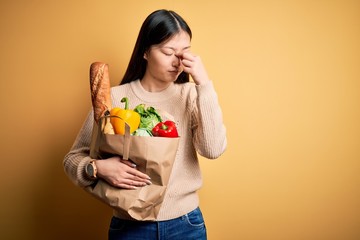 Young asian woman holding paper bag of fresh healthy groceries over yellow isolated background tired rubbing nose and eyes feeling fatigue and headache. Stress and frustration concept.