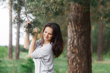 young attractive girl with long hair, brunette, in clothes of neutral colors, stands in the summer forest and holds a pine branch