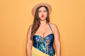 Young hispanic woman wearing summer hat and swimsuit over yellow background puffing cheeks with funny face. Mouth inflated with air, crazy expression.