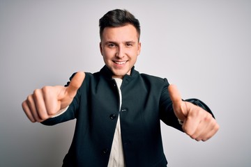 Young handsome business mas wearing elegant winter coat standing over isolated background approving doing positive gesture with hand, thumbs up smiling and happy for success. Winner gesture.