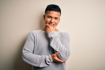 Young handsome latin man wearing casual sweater standing over isolated white background looking confident at the camera smiling with crossed arms and hand raised on chin. Thinking positive.