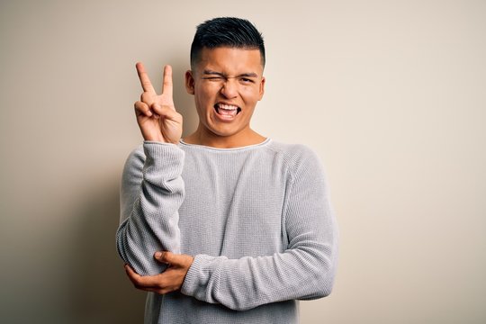 Young handsome latin man wearing casual sweater standing over isolated white background smiling with happy face winking at the camera doing victory sign. Number two.