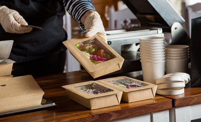 Restaurant worker wearing protective mask and gloves packing food boxed take away. Food delivery services and Online contactless food shopping.