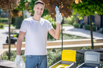 Smiling dark-haired male in protective gloves holding plastic packaging and empty bottle in front of waste container