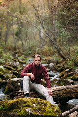bearded young white man in maroon hoody and milky jeans sitting on the old fallen mossy tree lying abroad the river in forest
