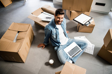Cheerful young man relaxing with laptop while moving to flat