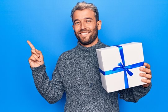 Young handsome blond man with beard holding birthday present over isolated blue background smiling happy pointing with hand and finger to the side