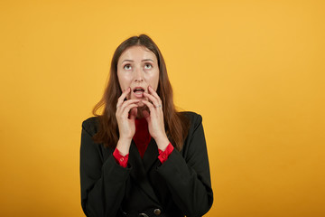 Young Attractive Brunette Woman In Black Stylish Suit, Red Shirt On Yellow Background, Confused Female Touches Hands To Her Mouth And Thinks. The Concept Of Uncertain, Changeable People, Indecisive