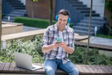 Cheerful dark-haired male wearing headphones, checking his smartphone, sitting on bench with his laptop