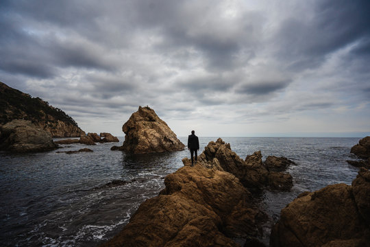 hombre de espalda parado sobre unas grandes rocas dentro del mar mediterr&aacute;neo con el cielo nublado