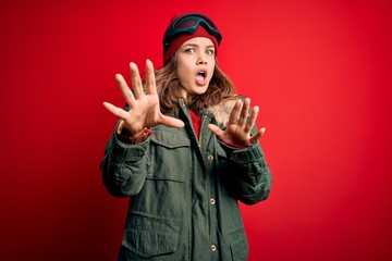 Young blonde girl wearing ski glasses and winter coat for ski weather over red background afraid and terrified with fear expression stop gesture with hands, shouting in shock. Panic concept.