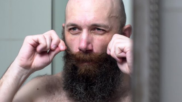 Bold man is combing his beard and mustache by hands. Attractive man in bathroom looks in mirror and combes black hair on his face.