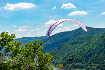 A man flying a white paraglider over the forest and the river on a beautiful sunny day.