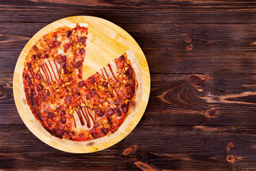 BBQ pizza with ham, bbq sauce, bacon and salami, without one slice on a wood platter which is on wooden rustic table, top view