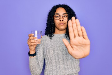 Young african american woman drinking glass of water over isolated purple background with open hand doing stop sign with serious and confident expression, defense gesture