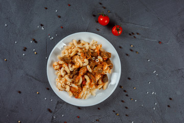 Pasta, macaroni with meat and sauce served on the white plate on the grey textured background. Background is decorated with salt, pepper and tomatoes. Delicious meal for lunch or dinner. 
