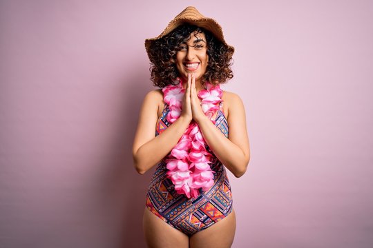 Young beautiful arab woman on vacation wearing swimsuit and hawaiian lei flowers praying with hands together asking for forgiveness smiling confident.