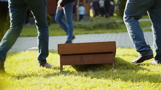 Men in trousers and jeans pick up bags of grains from cornhole and green grass in sunny weather