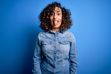 Young beautiful curly arab woman wearing casual denim shirt standing over blue background sticking tongue out happy with funny expression. Emotion concept.