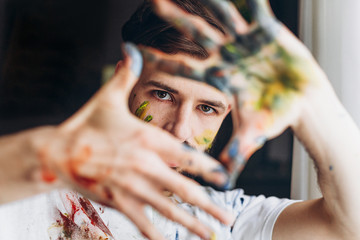 Portrait of a young creative artist, man wearing a white T-shirt with colored spots from paint posing for a photo in the process of creating a picture - obrazy, fototapety, plakaty