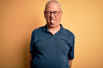 Middle age handsome hoary man wearing casual polo and glasses over yellow background sticking tongue out happy with funny expression. Emotion concept.