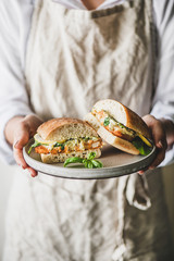 Woman in linen apron holding fresh fried fish sandwich with tartare sauce, lemon and arugula cut in...