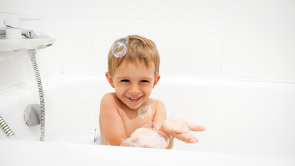 Portrait of cheerful laughing little boy washing in bath and cathing soap bubbles on hand