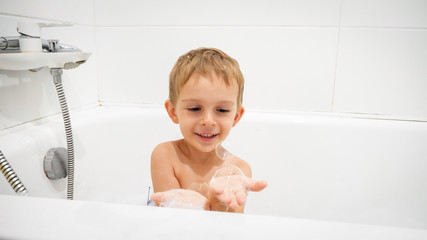 Portrait of cute little boy holding soap bubbles on hand while having bath