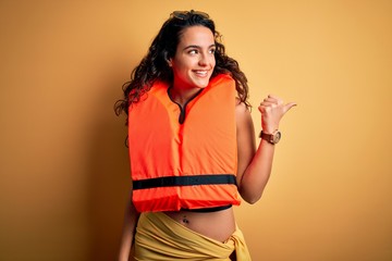 Young beautiful woman with curly hair wearing orange lifejacket over yellow background smiling with happy face looking and pointing to the side with thumb up.