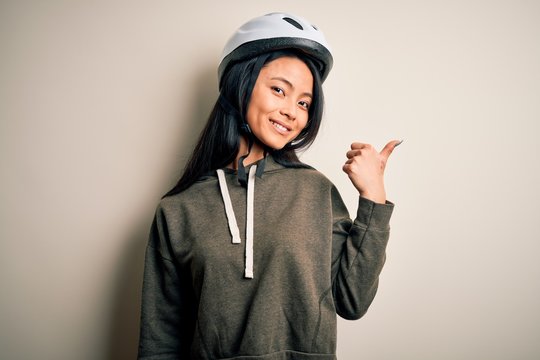 Young beautiful chinese woman wearing bike helmet over isolated white background smiling with happy face looking and pointing to the side with thumb up.