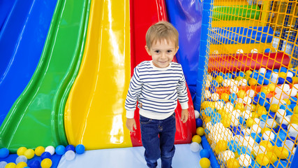 Fototapeta na wymiar Portrait of happy smiling little boy standing in playroom with colorful slide at amusement park