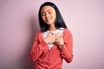 Young beautiful chinese woman wearing casual sweater over isolated pink background smiling with hands on chest with closed eyes and grateful gesture on face. Health concept.