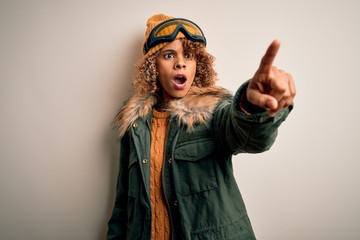 Young african american skier woman with curly hair wearing snow sportswear and ski goggles Pointing with finger surprised ahead, open mouth amazed expression, something on the front