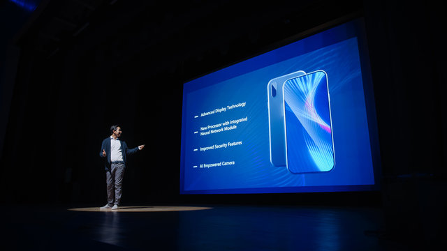 Live Event with Brand New Products Reveal: Speaker Presents Smartphone Device to Audience. Movie Theater Screen Shows Mock-up Touch Screen Mobile Phone with High-Tech Features and Top Highlights