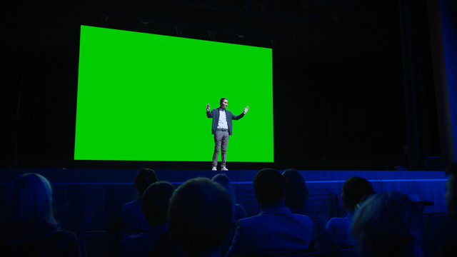 On Stage, Keynote Speaker Presents New Product to the Audience, Behind Him Movie Theater with Green Screen, Mock-up, Chroma Key. Business Live Event or Device Reveal