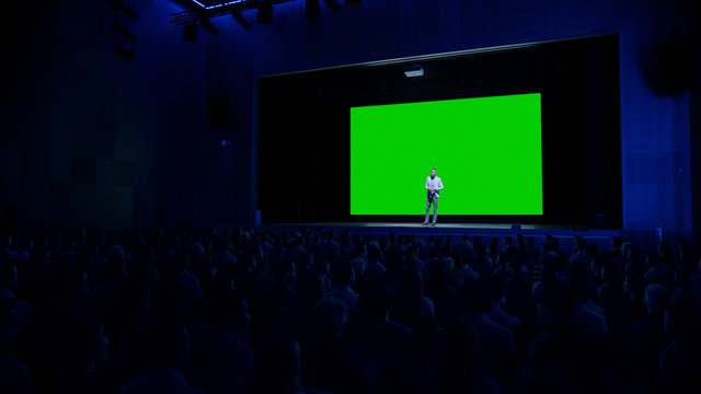 Keynote Speaker Does Presentation Of New Product To The Audience, Behind Him Movie Theater With Green Screen, Mock-up, Chroma Key. Business Conference Live Event Or Device Reveal