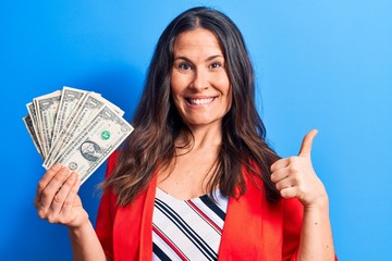 Beautiful brunette businesswoman holding bunch of dollars banknotes over blue background smiling happy and positive, thumb up doing excellent and approval sign