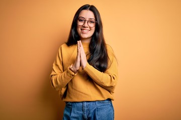 Young brunette woman wearing glasses and casual sweater over yellow isolated background praying with hands together asking for forgiveness smiling confident.