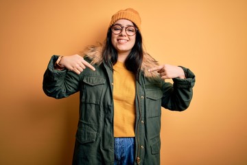 Young brunette woman wearing glasses and winter coat with hat over yellow isolated background...
