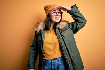 Young brunette woman wearing glasses and winter coat with hat over yellow isolated background very happy and smiling looking far away with hand over head. Searching concept.