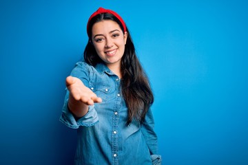Young brunette woman wearing casual denim shirt over blue isolated background smiling cheerful offering palm hand giving assistance and acceptance.